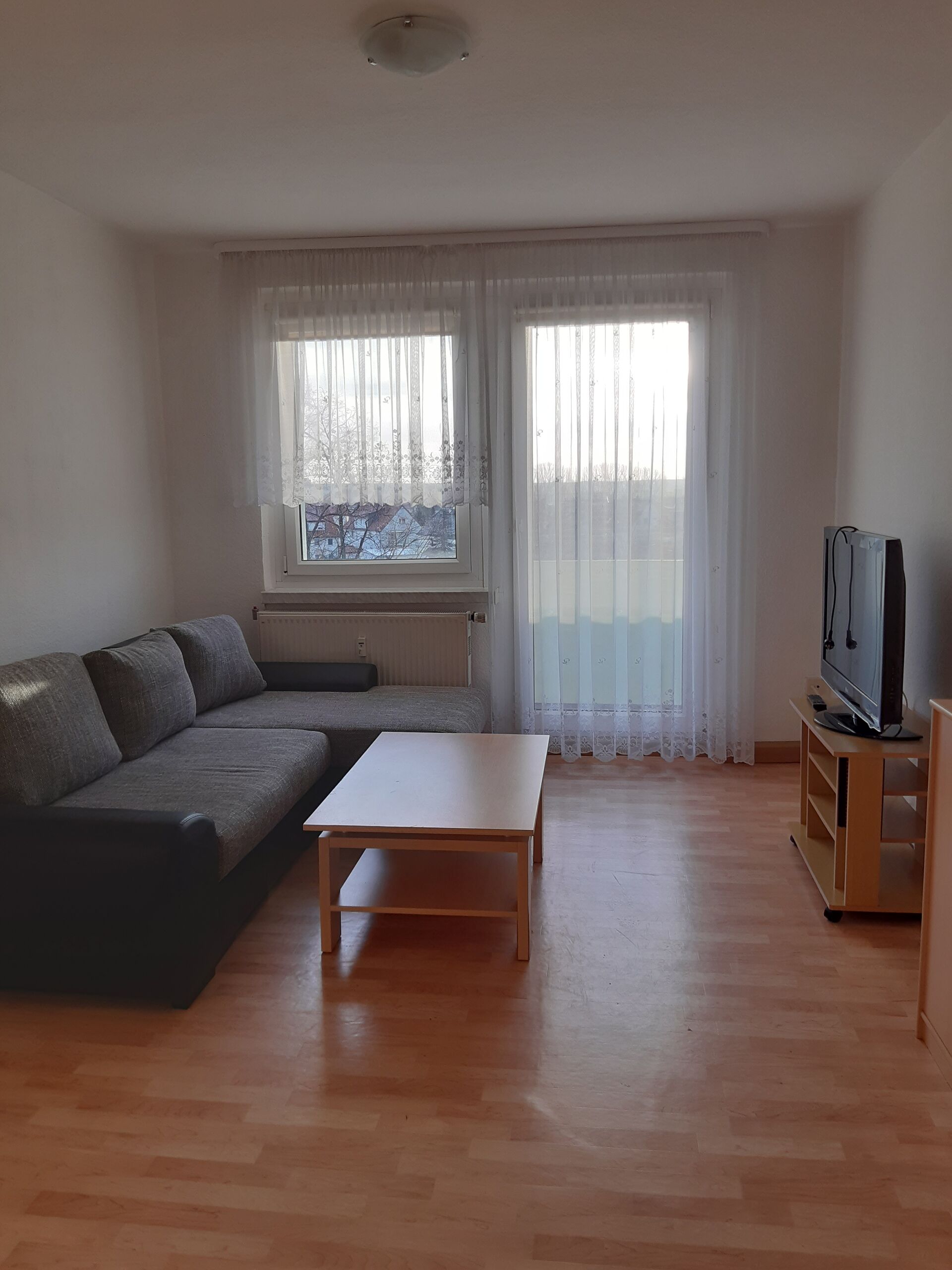 Gästewohnung in Gröditz, Am Kanal 25 - Wohnzimmer mit Fernseher, Holztisch, grauer Couch, Parkett und einem Balkon