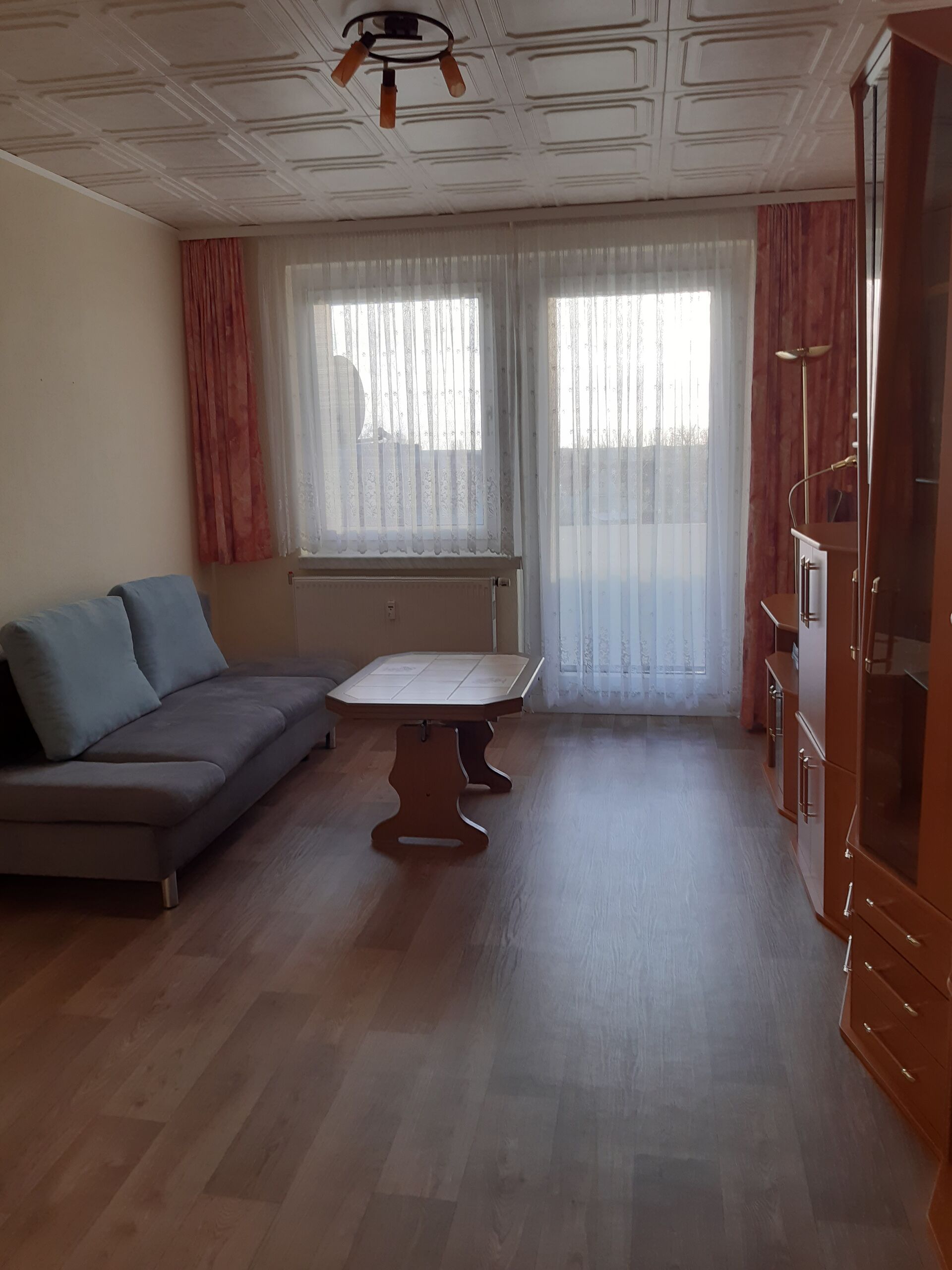Gästewohnung in Gröditz, Am Kanal 23 - Wohnzimmer mit Parkett, Holztisch, Couch und roten Gardinen