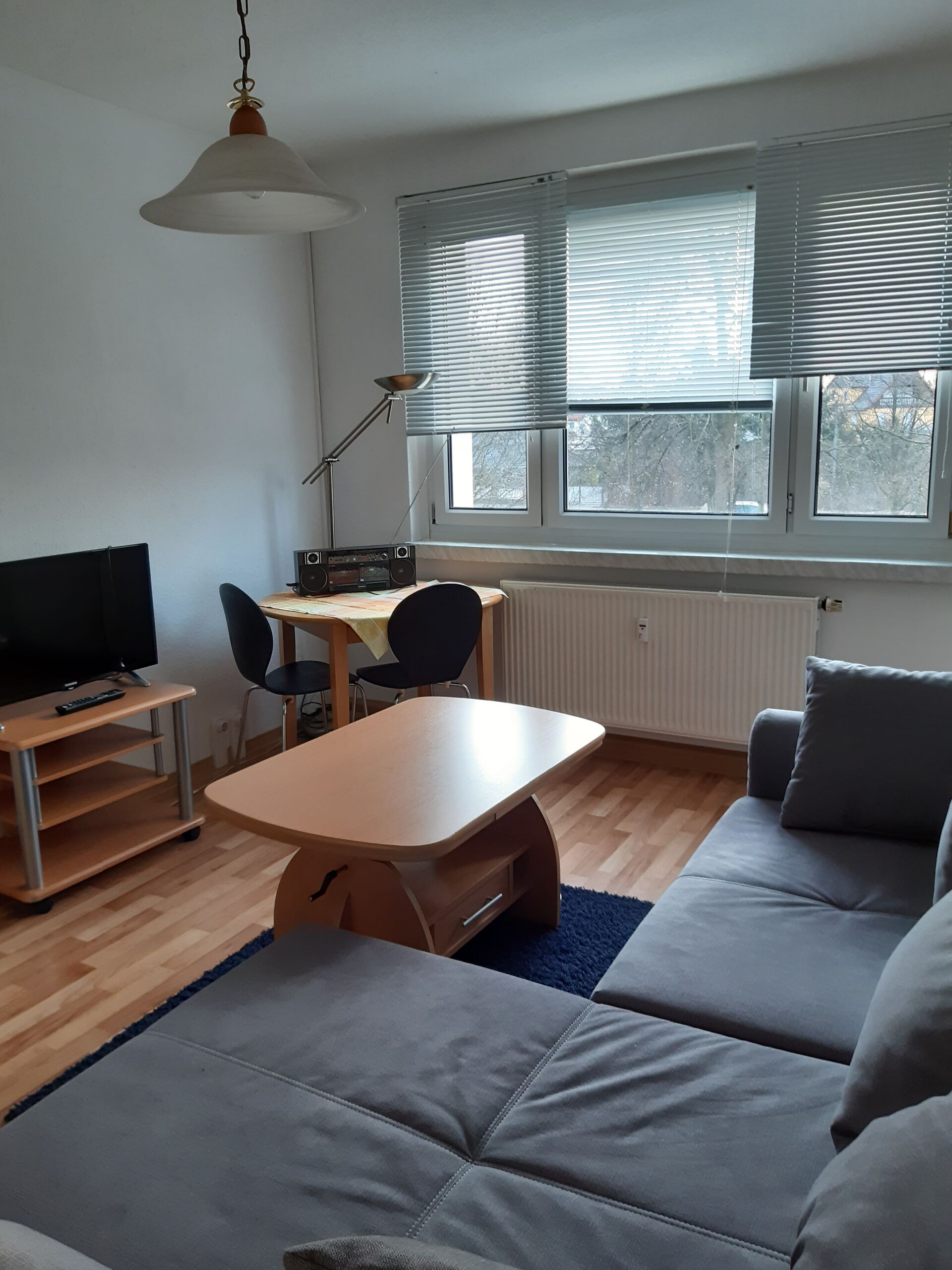 Gästewohnung in Gröditz, Am Kanal 27 - Wohnzimmer mit Parkett, Holztisch, Couch, Fernseher und Plissees